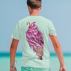 A person is wearing the Sideline Scoop Tee by Hobo Lax, a refreshing mint green T-shirt showcasing a vibrant ice cream cone graphic on the back. The design features pink ice cream with a cherry, beginning to melt. They're enjoying a beach day, bottle in hand, as ocean waves crash behind them—ideal attire for Summer 2024.