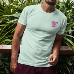 A man in a Hobo Lax Sideline Scoop Tee, featuring a refreshing mint color with pink writing, stands in front of lush green plants. He's wearing burgundy shorts and casually leaning on a wooden railing, perfectly capturing the laid-back vibe of Summer 2024.